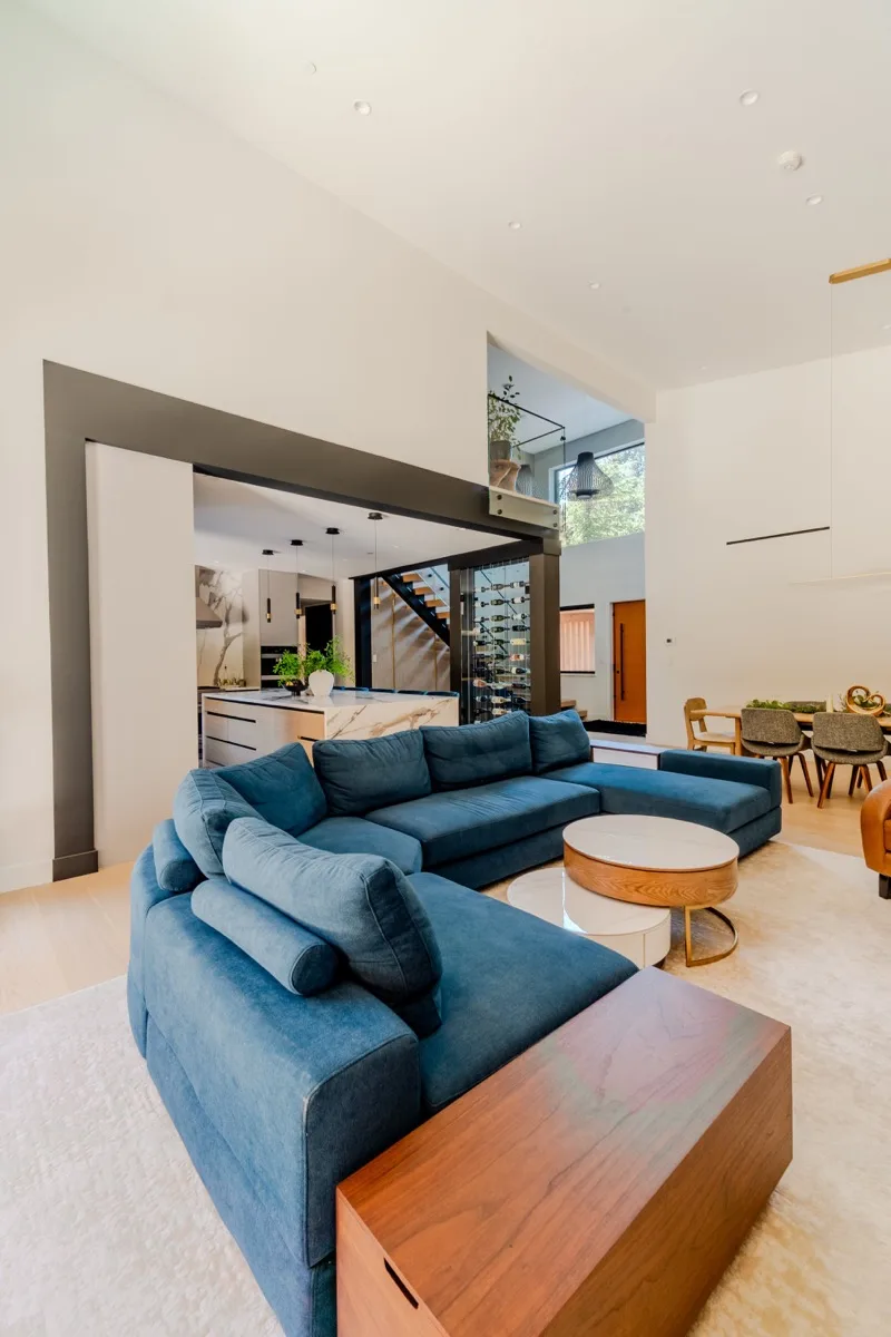 Open-plan living room real estate photography Vancouver, oversized blue velvet sectional, round gold coffee table, double-height ceilings, open-tread staircase, and open-plan kitchen beyond