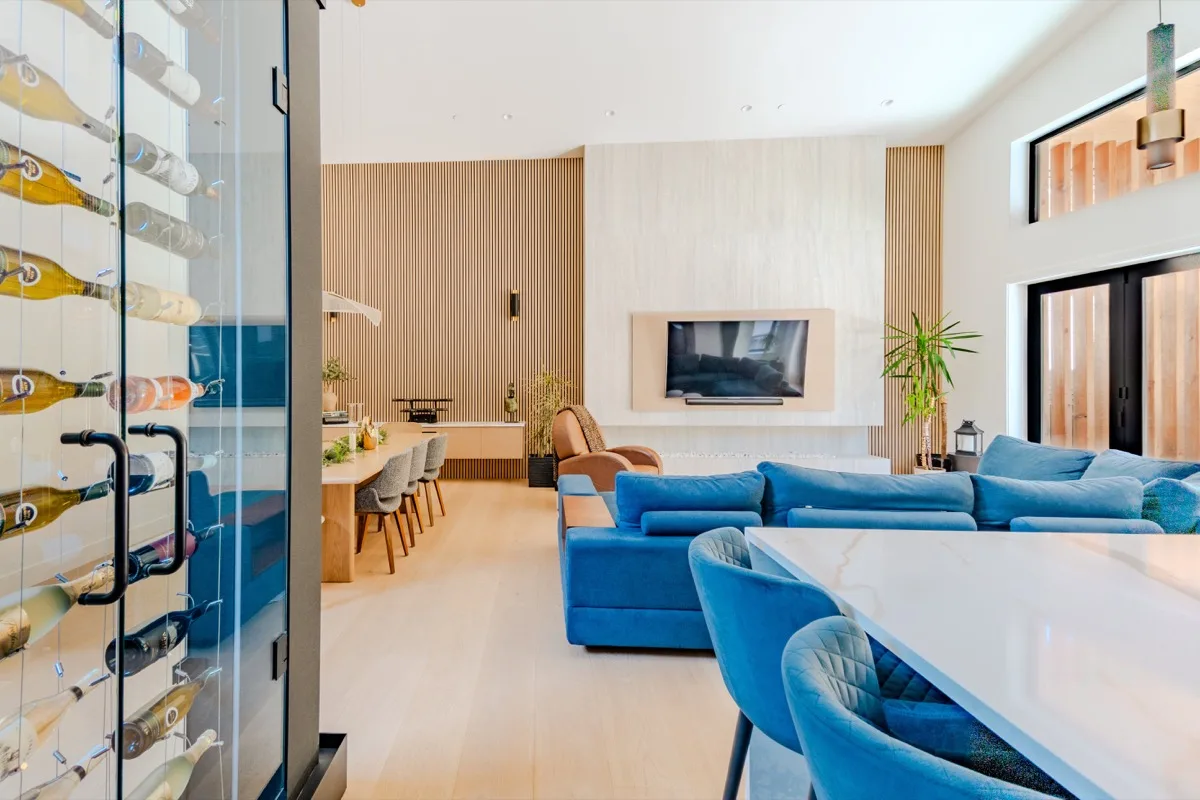 Open-plan living and dining real estate photography Vancouver, glass wine display wall, blue velvet sectional, marble dining table with velvet chairs, wood slat feature wall, and vaulted ceiling