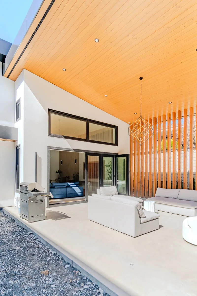 Covered outdoor patio real estate photography Vancouver, cedar tongue-and-groove ceiling, diamond geometric pendant, white lounge seating, glass sliding doors opening to interior, and vertical wood slat privacy screen