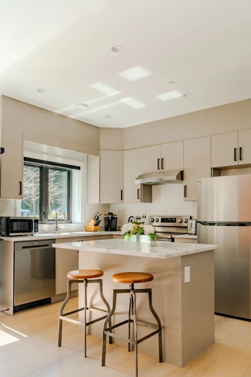 Kitchen real estate photography Vancouver, taupe flat-front cabinetry, quartz island with industrial wood and metal bar stools, stainless steel appliances, skylights, and garden window