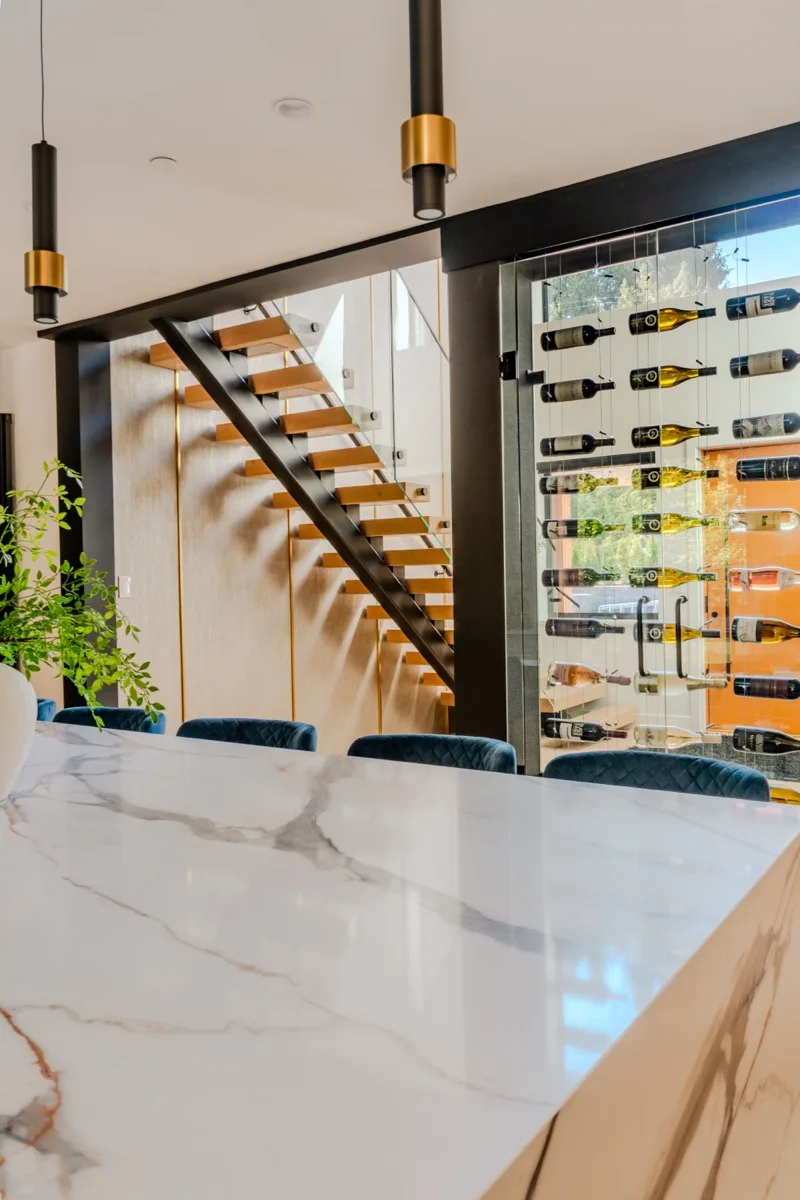 Dining area real estate photography Vancouver, white marble waterfall island, floor-to-ceiling glass wine display wall, floating staircase with brass cylinder pendants, and teal velvet dining chairs